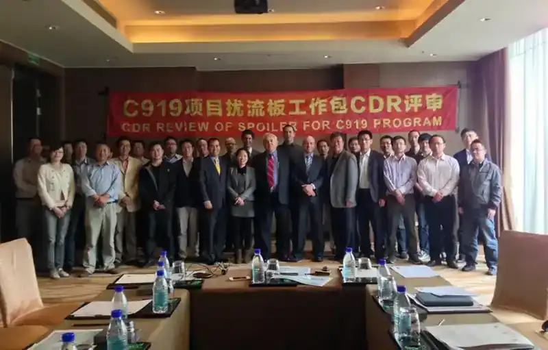 Full international joint engineering team gathered for the C919 Spoiler Critical Design Review — COMAC and partner engineers in front of bilingual programme banner, group photo around conference table