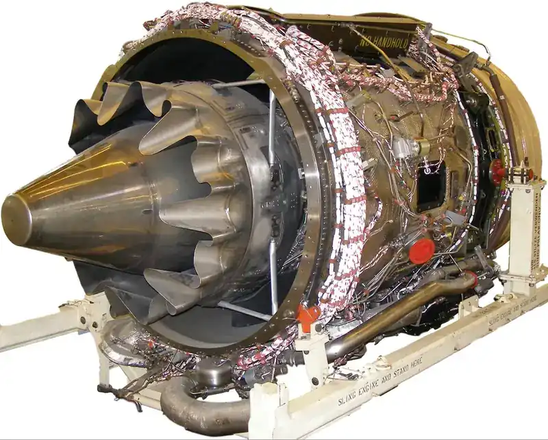 Rolls-Royce BR710 high-bypass turbofan engine on a transport stand — exhaust end view showing the characteristic forced-mixer chevron nozzle, low-pressure turbine outer casing wrapped with instrumentation wiring harnesses, and accessory pipework. On display at Tempelhof.