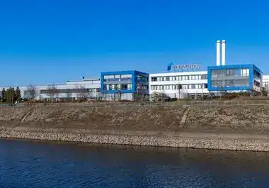 Rheinmetall facility in Neuss, Germany — blue-and-white office and production buildings on the riverbank, viewed across the water on a clear winter day