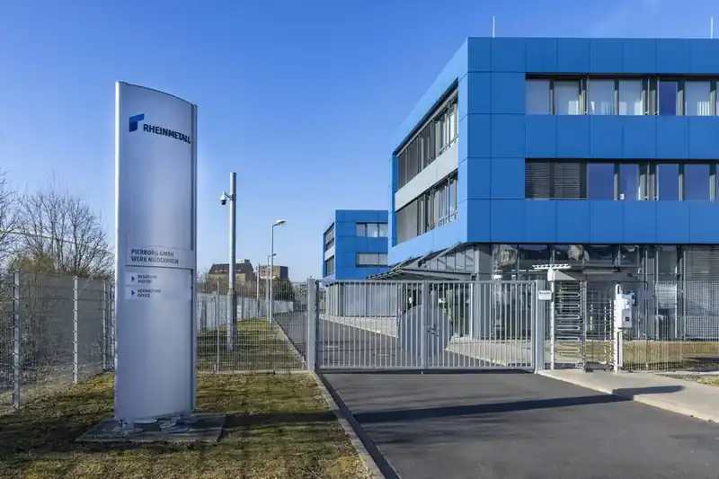 Pierburg GmbH Werk Niederrhein (a Rheinmetall company) entrance gate — visitor signage, blue-cladded office buildings, perimeter fencing and turnstiles on a bright winter day