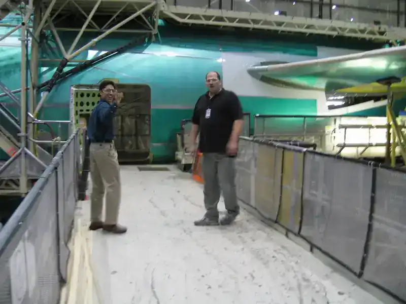 Two engineers on the Boeing factory floor wearing safety glasses, with a green-primed 737 fuselage section visible in the background