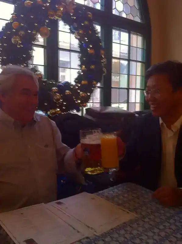 Steve Clamp toasting with Dr Shiu, COMAC C919 Design Manager, in Munich, Germany during the C919 development phase — Bavarian beer hall setting with Christmas decorations and traditional stained-glass window