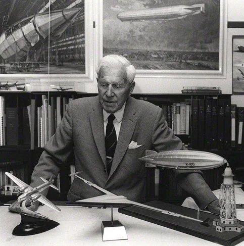 Airship pioneer at his desk with airship paintings and an R-100 model behind him