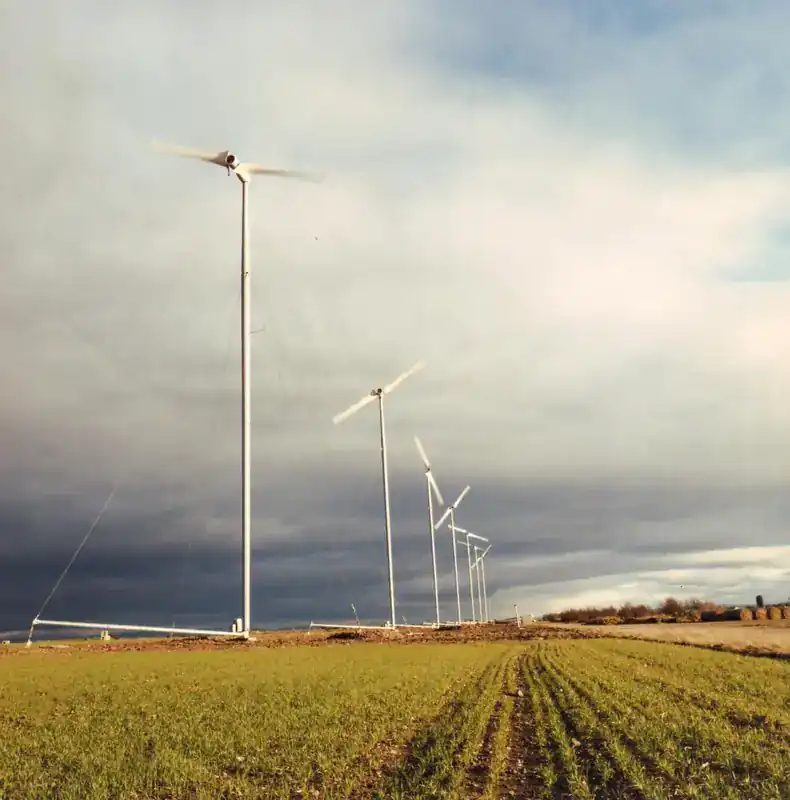 Carter Model-300 wind turbines installed in a row at Great Orton wind farm in Cumbria, England — guyed-tower, two-bladed teetering hub configuration on a freshly seeded field with dramatic cloud-covered sky