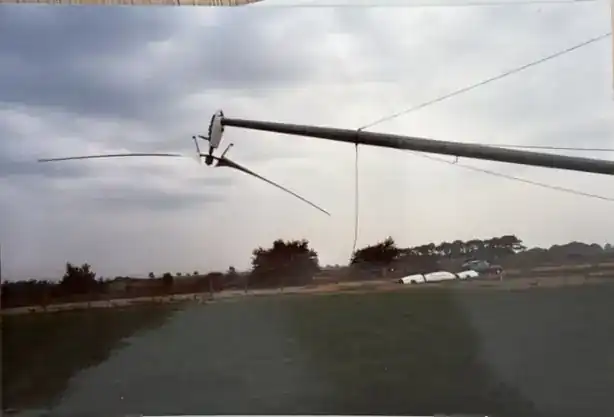 Carter wind turbine in lowered/tilted position — two-bladed teetering rotor and nacelle suspended low, supported by guy cables. Single guyed-tower turbine seen from ground level with hedgerow and cars in background.