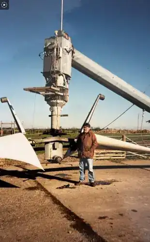 Engineer (Steve Clamp) stood beside a Carter wind turbine nacelle and rotor assembly that has been lowered to ground level for maintenance — close-up view showing the gearbox, generator and hub mechanism, with one blade visible on the ground