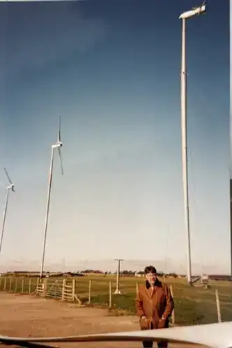 Engineer stood beside three Carter wind turbines in a row — guyed-tower, two-bladed teetering hub configuration, clear blue sky, ground-level perspective showing tower bases and guy-wire anchors